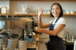 Enthusiastic asian barista, girl using coffee machine, waving at client, saying hello, greeting guests in her cafe photo