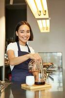 Vertical shot of smiling girl working as barista, prepare pour over, making filter coffee batch brew, standing at counter in cafe in apron photo