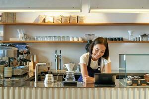 Happy smiling asian barista, girl behind counter, working with POS terminal and brewing filter kit, making coffee in cafe photo