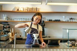 Smiling asian girl barista, pouring hot water from kettle, brewing filter coffee, standing behind counter in cafe photo