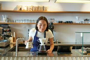 Happy smiling asian girl smelling coffee while brewing filter, standing behind counter in cafe photo