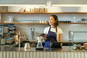 retrato de sonriente asiático niña barista, en pie cerca café fabricación de cerveza equipo, haciendo filtrar en cafetería, participación pos Terminal, Procesando clientela orden foto