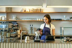 retrato de linda niña estudiante trabajos como barista, sostiene pos crédito tarjeta máquina, en pie a mostrador con terminal y fabricación de cerveza equipo foto