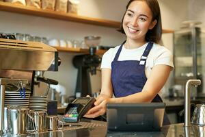 Smiling asian girl barista, giving client POS terminal, help pay with credit card, standing at counter in coffee shop, working in cafe photo