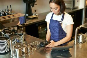 sonriente asiático niña barista trabajando en café a encimera, Procesando pedidos, utilizando pos terminal recepción pago para café foto