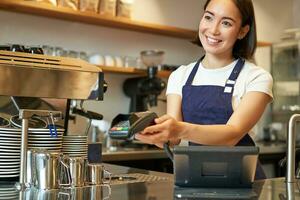Smiling asian girl barista, giving client POS terminal, help pay with credit card, standing at counter in coffee shop, working in cafe photo