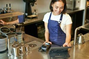 sonriente asiático niña barista trabajando en café a encimera, Procesando pedidos, utilizando pos terminal recepción pago para café foto