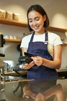 hermosa sonriente asiático chica, barista insertar cliente crédito tarjeta en pos Terminal, Procesando pago, tomando pedidos en café tienda foto