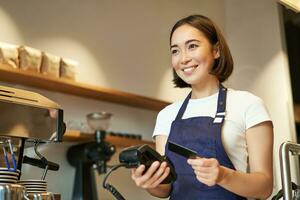 linda asiático chica, barista en café Procesando sin contacto pago, insertar crédito tarjeta dentro pos Terminal, tomando orden en café tienda foto