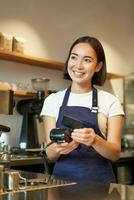 retrato de sonriente asiático barista, café tienda empleado utilizando pos terminal y crédito tarjeta, ayuda cliente pagar sin contacto en café foto