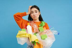 Woman with complicated face, looking confused at plastic garbage, clueless how to recycle items, go green, standing over blue background isolated photo