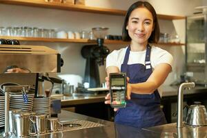 sonriente asiático barista, café tienda personal da usted crédito tarjeta máquina, Procesando pago con pos Terminal, trabajando en café foto