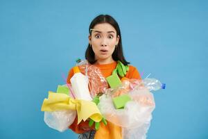 Young confused asian girl with different types of plastic, learns how to recycle, saving enviroment, sorting garbage, blue background photo