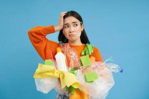Woman with complicated face, looking confused at plastic garbage, clueless how to recycle items, go green, standing over blue background isolated photo