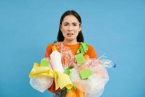 ceñudo asiático mujer con el plastico basura en manos, quejumbroso a no reciclaje desperdiciar, en pie con camada terminado azul antecedentes foto