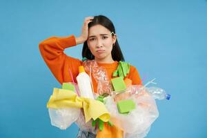 Woman with complicated face, looking confused at plastic garbage, clueless how to recycle items, go green, standing over blue background isolated photo