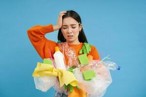 Woman with complicated face, looking confused at plastic garbage, clueless how to recycle items, go green, standing over blue background isolated photo