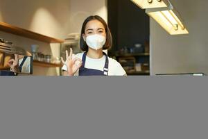 Asian female barista, girl in medical mask, shows okay sign, works in cafe behind counter, brews coffee, works with clients during covid pandemic photo