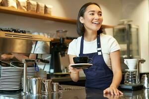 Portrait of beautiful asian girl, student working part-time in cafe, holding cup of coffee, made order, looking for client, wearing uniform photo