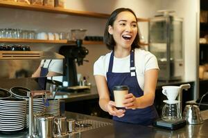 retrato de sonriente asiático niña barista, dando fuera orden en cafetería, atractivo invitado a recoger arriba para llevar orden cerca encimera, participación para llevar taza de café foto