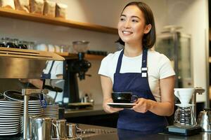 Portrait of smiling young woman providing customer service in cafe, holding cup of coffee, giving order to client, wearing apron uniform photo