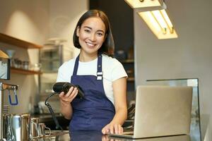 Smiling asian barista, girl with card terminal, payment machine and laptop, standing in cafe, processing payment for coffee order photo