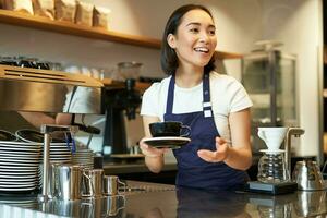 Portrait of beautiful asian girl, student working part-time in cafe, holding cup of coffee, made order, looking for client, wearing uniform photo