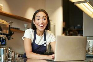 entusiasta asiático niña en café uniforme, barista trabajador con computadora portátil, mirando contento y sorprendido a cámara foto