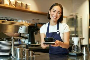 Portrait of beautiful asian girl, student working part-time in cafe, holding cup of coffee, made order, looking for client, wearing uniform photo