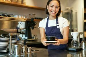 Portrait of smiling young woman providing customer service in cafe, holding cup of coffee, giving order to client, wearing apron uniform photo