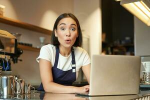 Portrait of asian girl, barista in apron with laptop, standing at counter in coffee shop, looking curious and surprised at camera photo