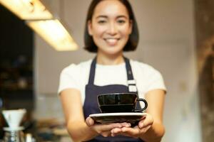retrato de sonriente asiático niña barista, camarera en café uniforme, dando usted taza de café, preparar bebida para cliente, mirando simpático foto