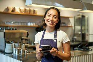 Portrait of smiling asian woman holds cup of coffee, prepare drinks for clients in cafe, working and serving drinks, wearing uniform apron photo