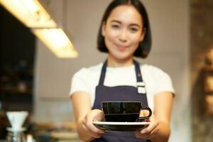 sonriente asiático niña barista, en pie en delantal uniforme, dando usted taza de café, hecho un bebida para cliente en café foto