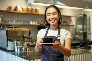 Portrait of smiling asian woman holds cup of coffee, prepare drinks for clients in cafe, working and serving drinks, wearing uniform apron photo