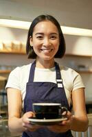 Vertical shot of friendly asian girl smiling, serving coffee, barista giving you cup of coffee, prepared drink for client in cafe photo