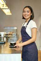 Vertical shot of smiling asian girl barista, wearing uniform, making coffee, standing near counter with cup, working in cafe photo