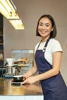 Vertical shot of smiling asian girl barista, wearing uniform, making coffee, standing near counter with cup, working in cafe photo