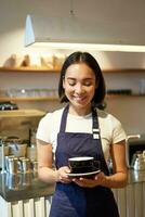 Vertical shot of friendly asian girl smiling, serving coffee, barista giving you cup of coffee, prepared drink for client in cafe photo