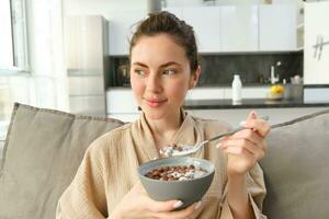 relajante Mañana a hogar. joven hermosa mujer en bata de baño, comiendo cereales en sofá, teniendo su desayuno en vivo habitación foto