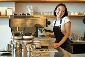 contento sonriente barista chica, coreano barman en delantal, en pie cerca café máquina, preparar orden para café cliente, mirando alegre a cámara foto