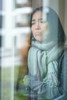 Portrait of sad ill korean woman looking out of the window, staying at home due to coronavirus, being sick and cant leave house photo