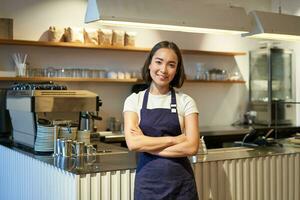 Smiling young barista wearing apron, working in cafe, standing near counter with coffee machine, looking friendly, inviting people for drink photo