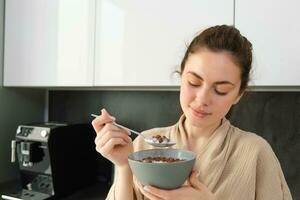 maravilloso mujer empieza su día con cereales, come desayuno y sonrisas, usa bata de baño, se inclina en cocina encimera, empieza su Mañana con rápido comida foto