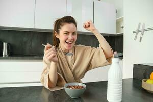 Portrait of happy, laughing young woman eating cereals with milk, triumphing, having breakfast and feeling excited, energetic morning concept photo