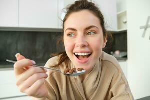 cerca arriba retrato de contento sonriente morena en bata de baño, participación cereales en cuchara, comiendo desayuno con Leche y mirando alegre, comenzando su día en Mañana foto