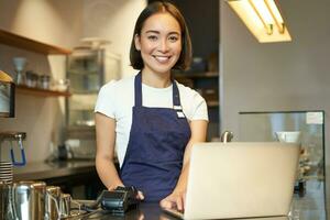 Smiling asian girl barista, working in cafe, serving client, looking at laptop while using card reader to receive money for order, selling coffee photo