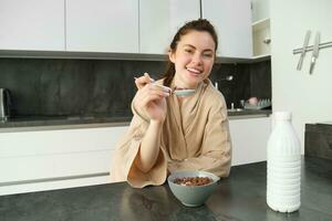 Portrait of happy young woman leans on kitchen worktop and eating cereals, has milk and bowl in front of her, having her breakfast, wearing bathrobe photo