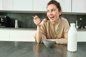 Portrait of young beautiful woman in bathrobe, eating cereals for breakfast, leans on kitchen worktop, looking at her morning meal photo