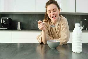 Portrait of beautiful young and healthy woman in bathrobe eats her breakfast in kitchen, has cereals with milk and smiling photo
